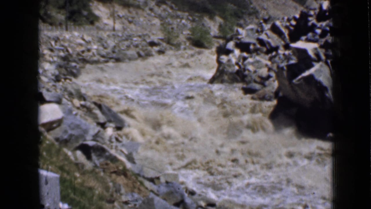 Vintage 8mm Utah frame showing a churning muddy river beside boulders, dry canyon ground, and sparse brush.