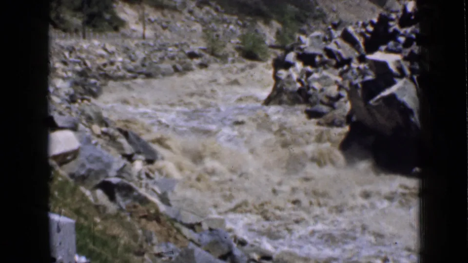 Vintage 8mm Utah frame showing a churning muddy river beside boulders, dry canyon ground, and sparse brush.