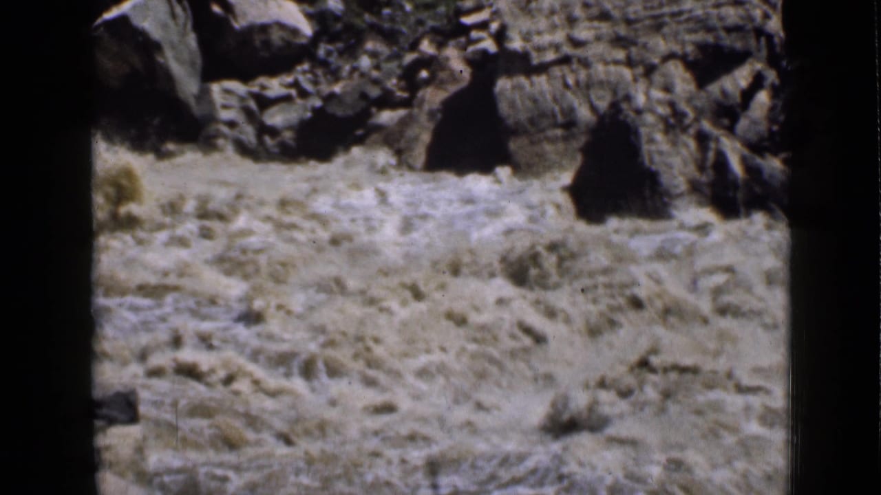 1939 Utah 8mm frame with muddy water surging below dark canyon rocks and scattered dry vegetation.