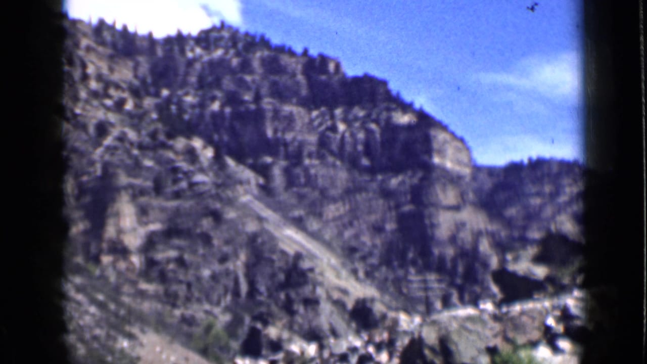 Rocky canyon slope with evergreen trees beneath a bright blue sky.