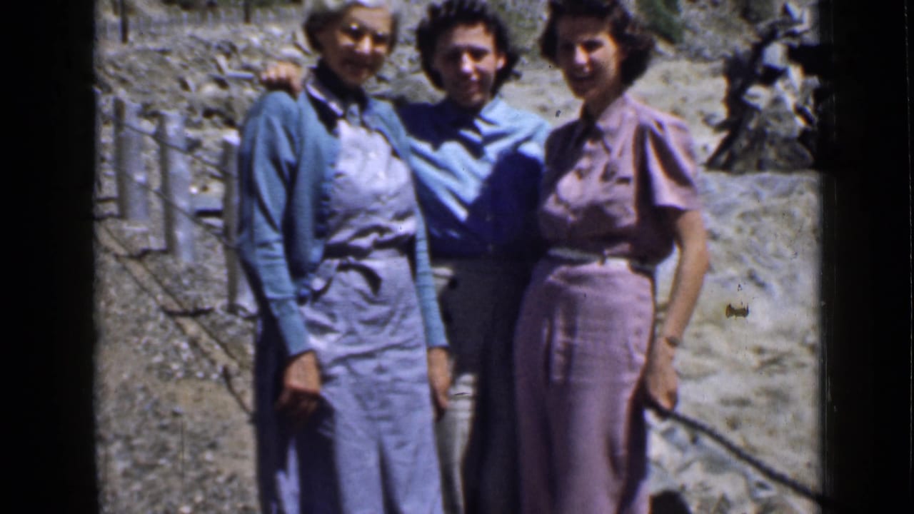 A home-movie frame shows three women on a rocky Utah hillside, smiling beside a rope or rail in mountain terrain.