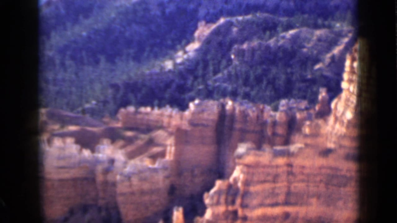 1939 Utah home-movie frame with orange sandstone walls in the foreground and dark wooded hills rising behind.