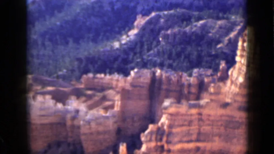 1939 Utah home-movie frame with orange sandstone walls in the foreground and dark wooded hills rising behind.