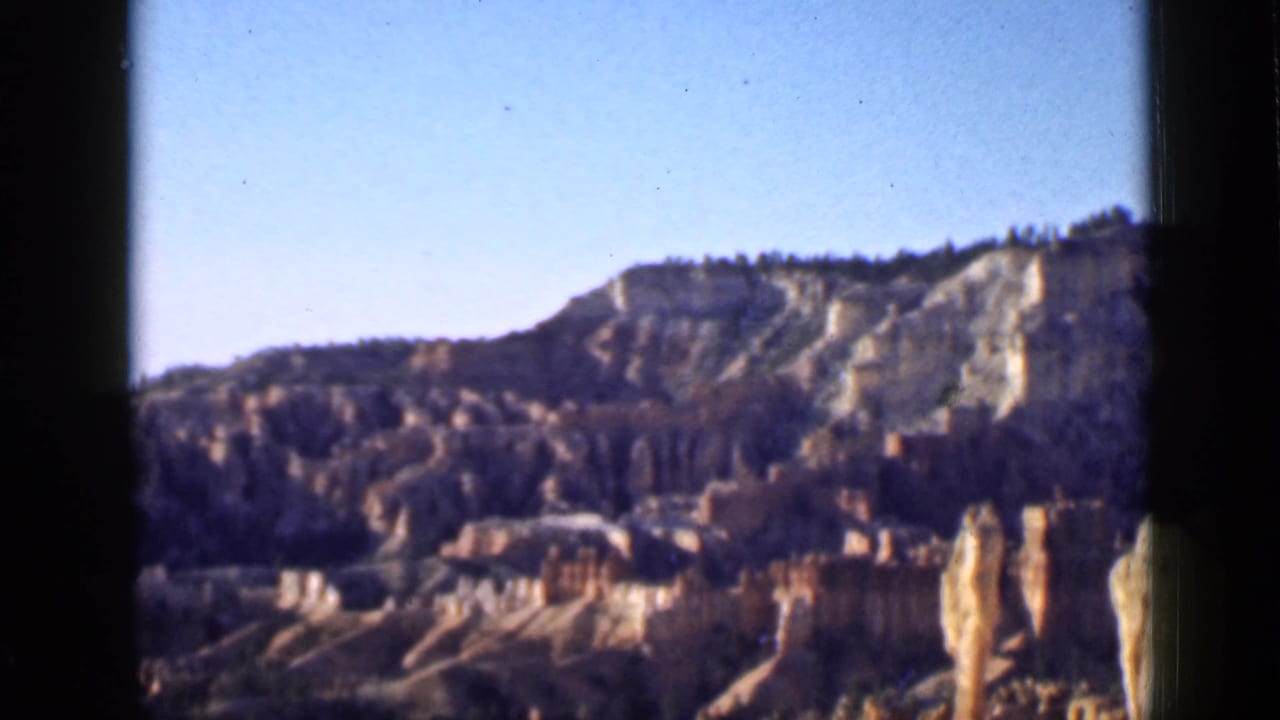 1939 Utah home-movie frame showing a wider canyon vista with blue sky, pale cliff bands, red spires, and dark vegetation.