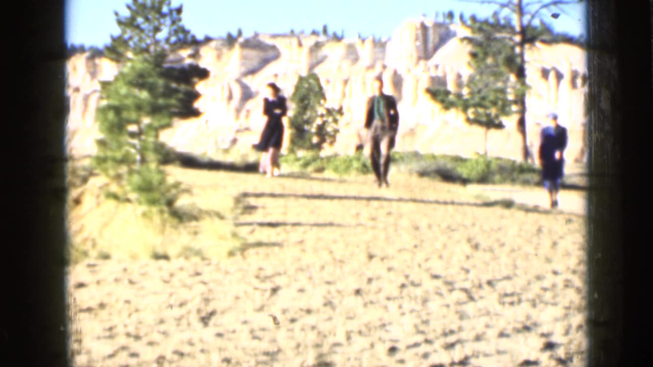 Three people walk on a dirt path near pale rock formations and pine trees.