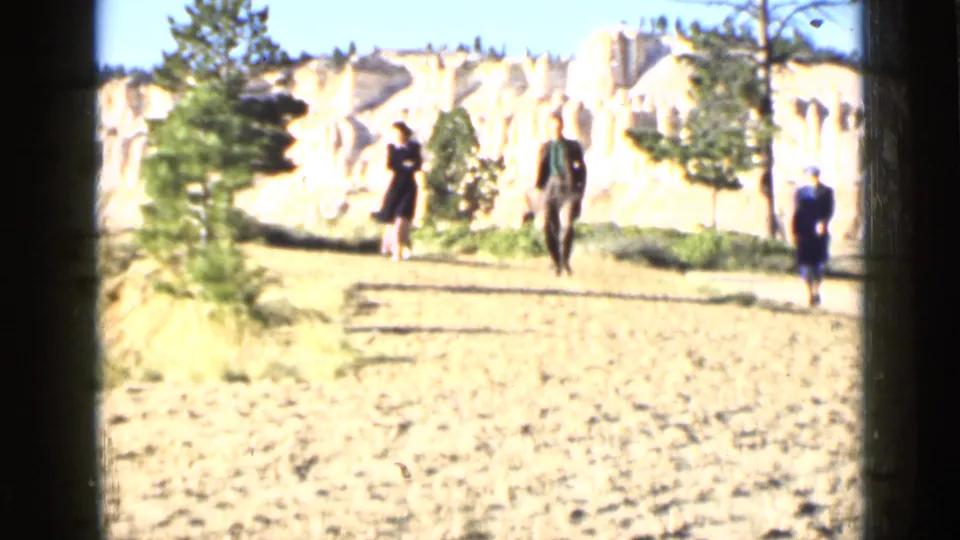 Three visitors walk along a dirt path in a Utah rock landscape, with pale formations and pine trees behind them.