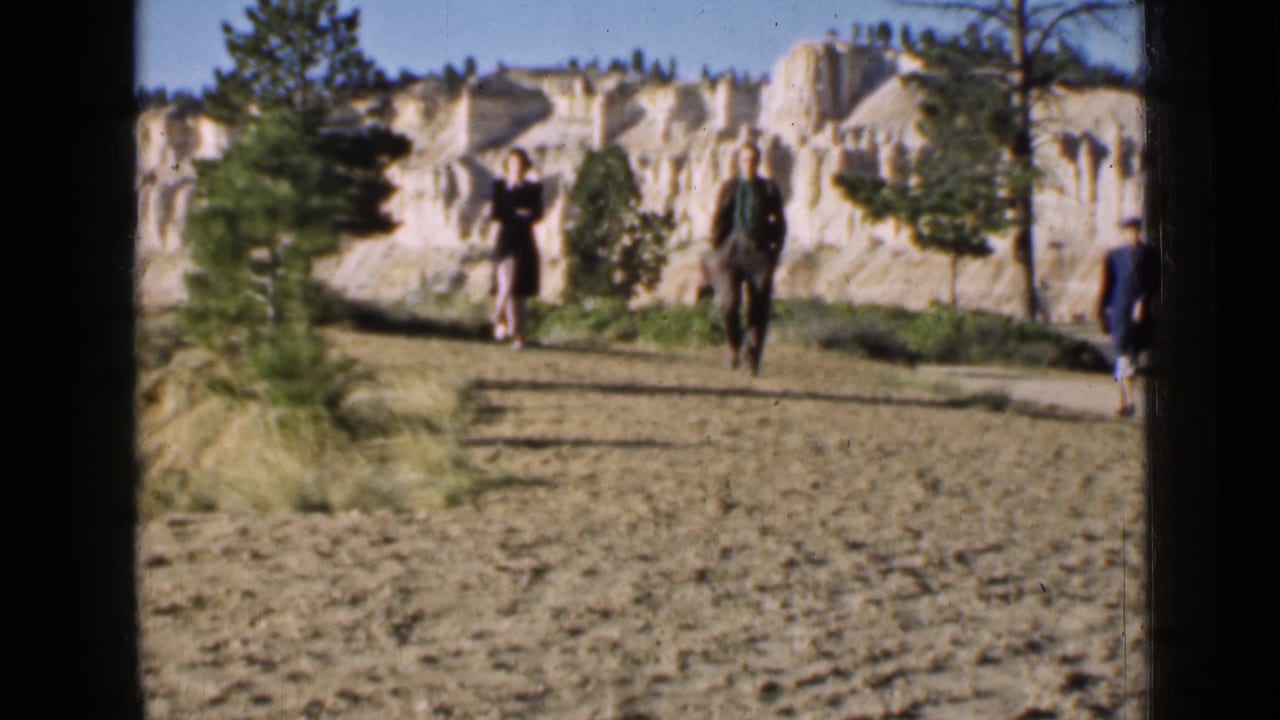 A suited man and a woman in a dress appear to walk toward the camera on a sandy Utah path, with rock formations behind.
