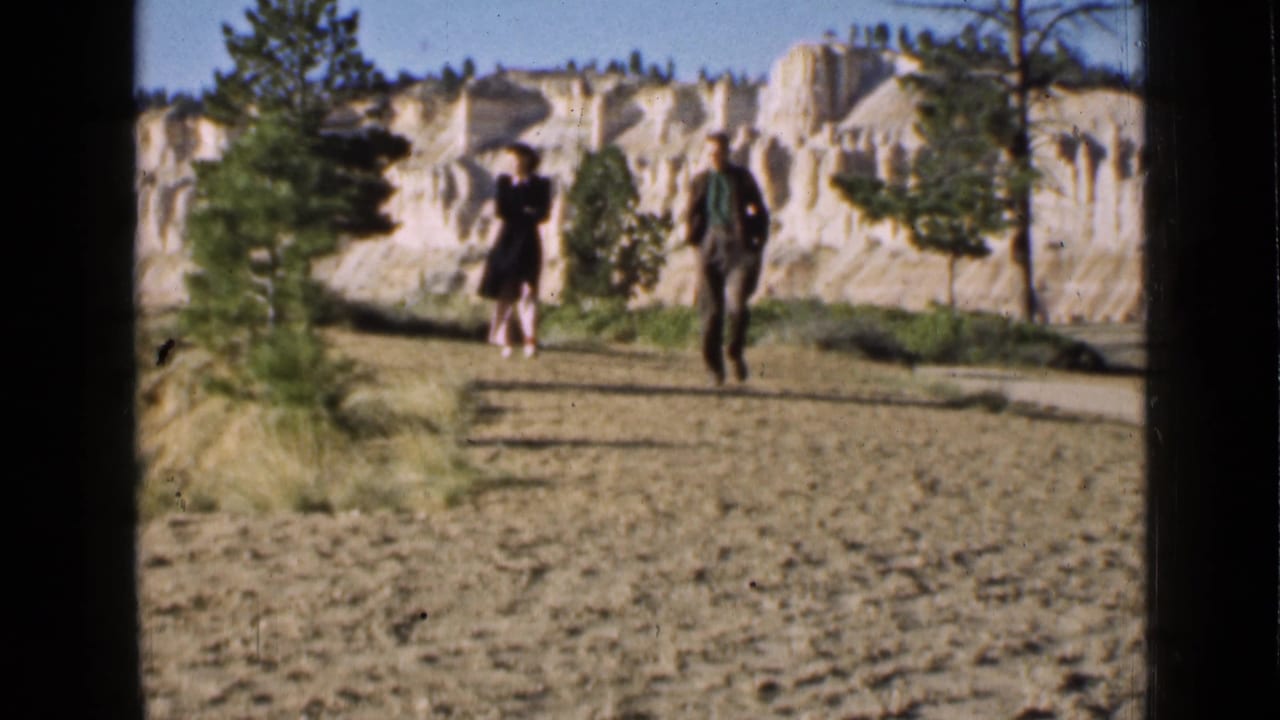 Two visitors remain centered on the path in this 1939 Utah travel frame, backed by pale eroded rock and sparse trees.
