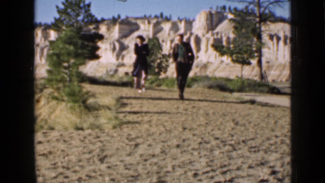 Two adults walking on a dirt path below pale cliffs and pine trees.