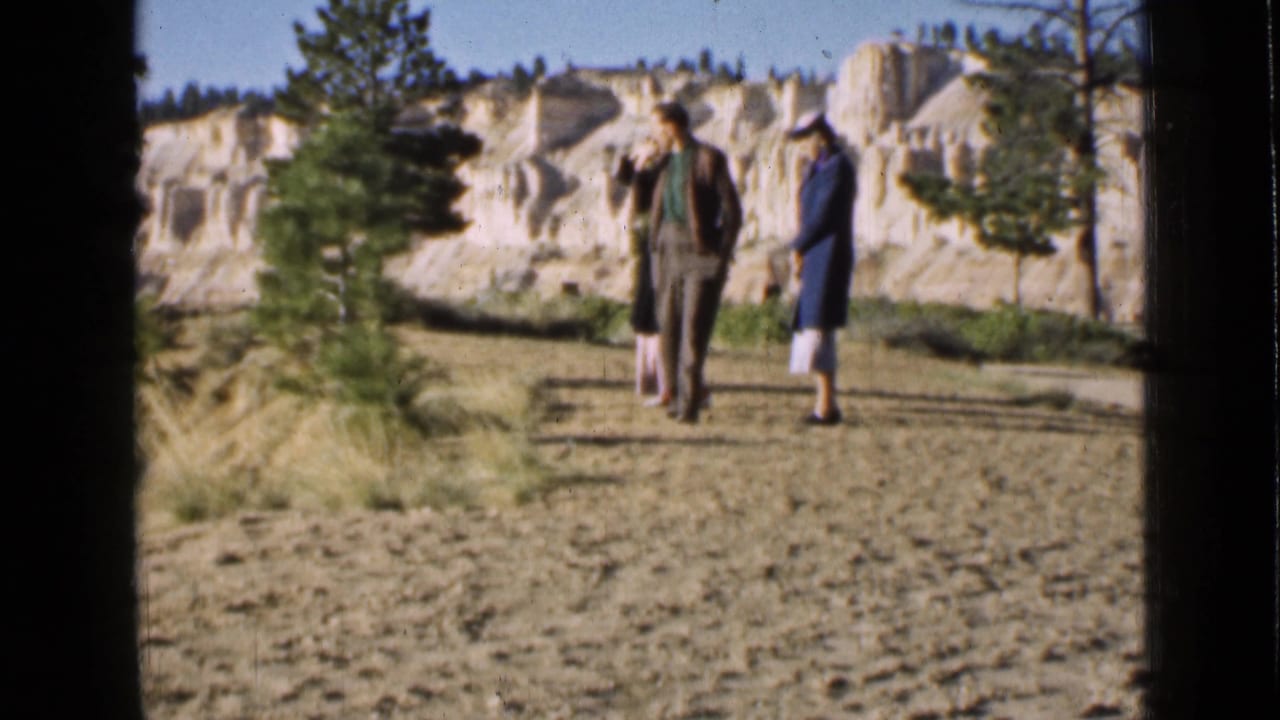 Three adult visitors walk away from the camera on a dry Utah park path, framed by pines and pale eroded cliffs.