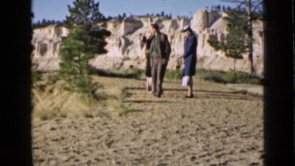 Three adult visitors walk away from the camera on a dry Utah park path, framed by pines and pale eroded cliffs.