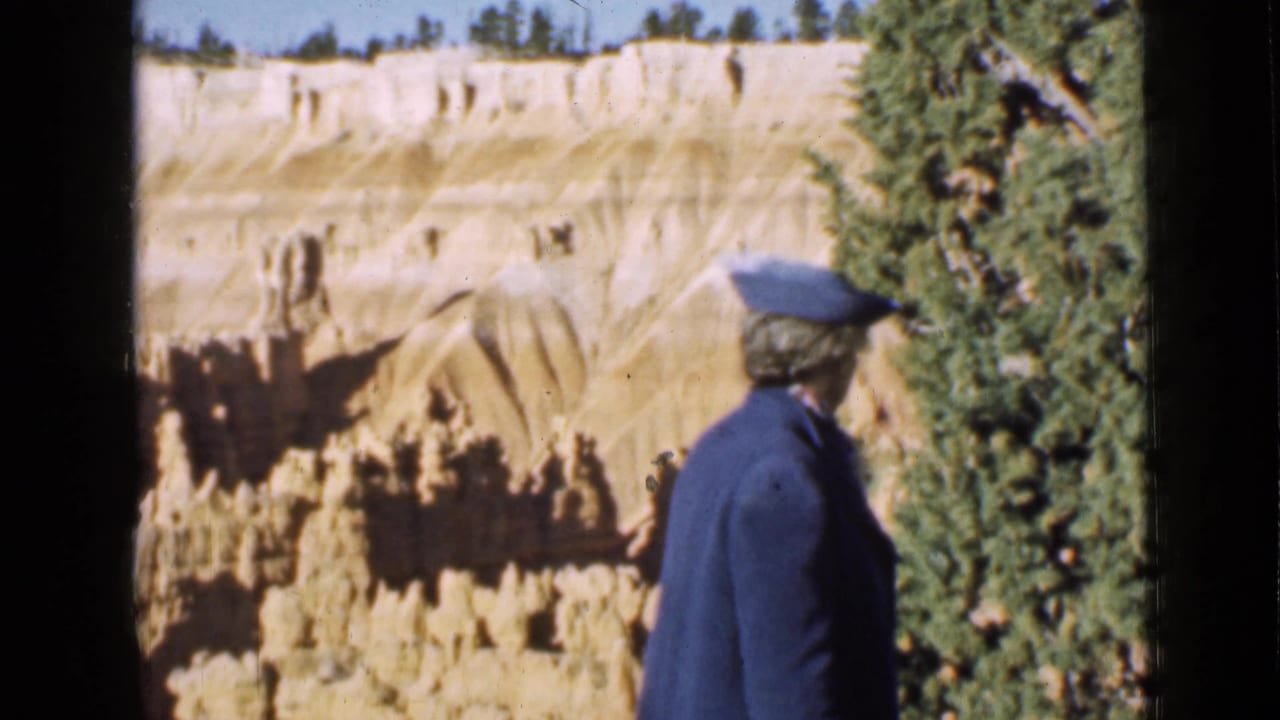 In Utah 1939 footage, a woman in a blue coat and hat looks toward Bryce Canyon hoodoo formations near a pine tree.