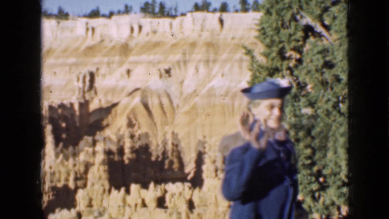 Woman in a blue coat and hat smiles and waves with canyon formations behind her.