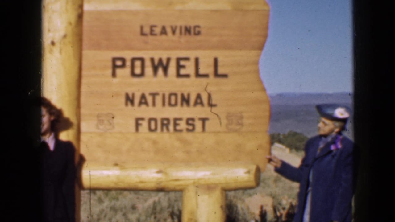 A wooden roadside sign reading LEAVING POWELL NATIONAL FOREST fills the frame, with two adults partly visible at the edges.