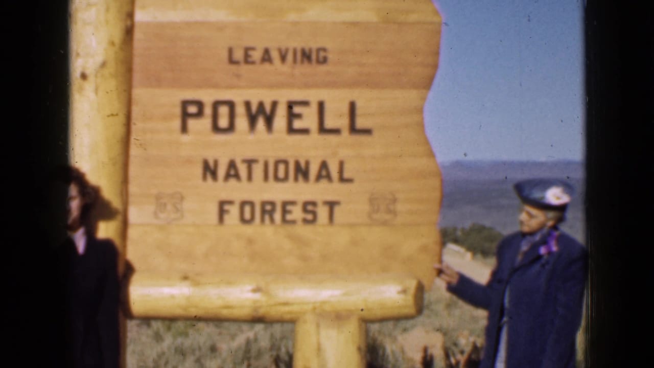 The woman in blue stands beside and points toward the LEAVING POWELL NATIONAL FOREST sign in 1939 Utah travel footage.