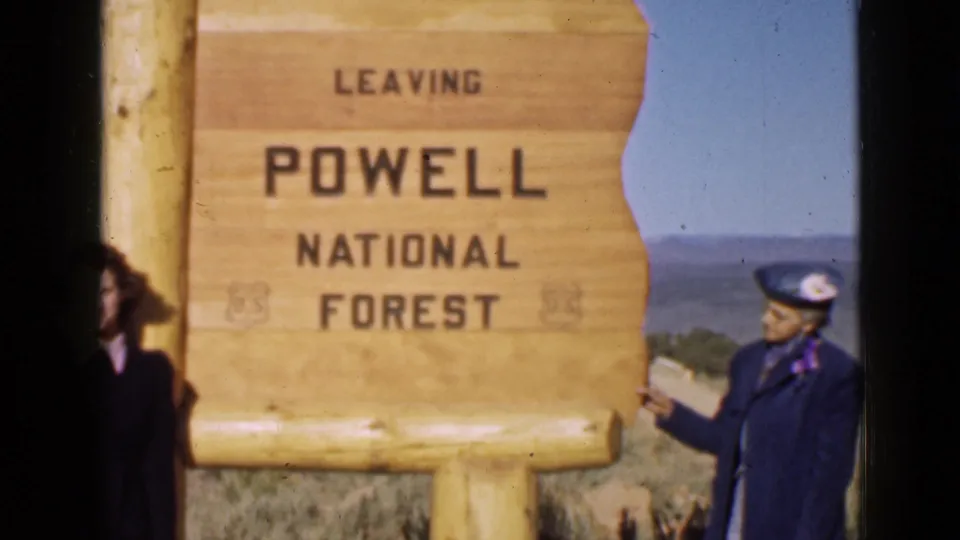 The woman in blue stands beside and points toward the LEAVING POWELL NATIONAL FOREST sign in 1939 Utah travel footage.