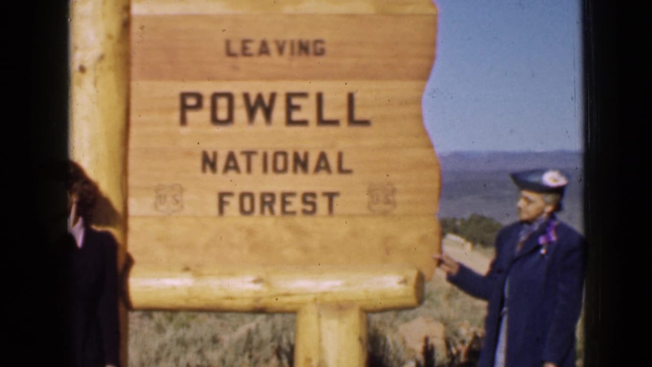1939 Utah home-movie frame showing adults near a Powell National Forest sign, with arid hills in the background.