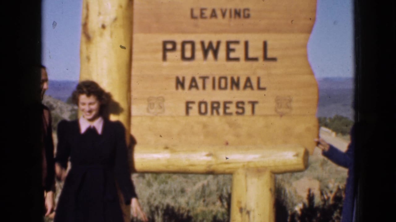 1939 Utah home-movie frame with a smiling woman standing beside the Powell National Forest sign.
