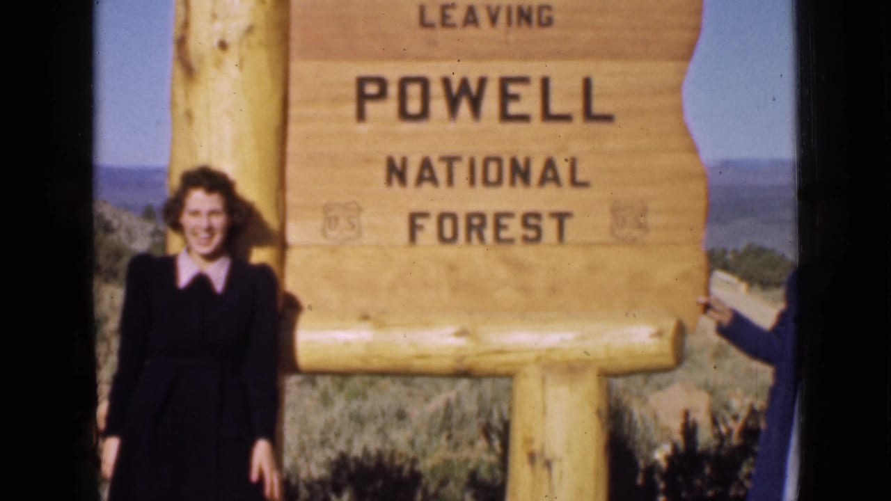 1939 Utah frame showing a woman posing by the Powell National Forest sign, with open hills and sky behind.