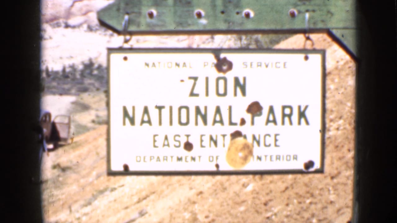 A National Park Service sign reads Zion National Park East Entrance, with a sandy road and parked car partly visible nearby.