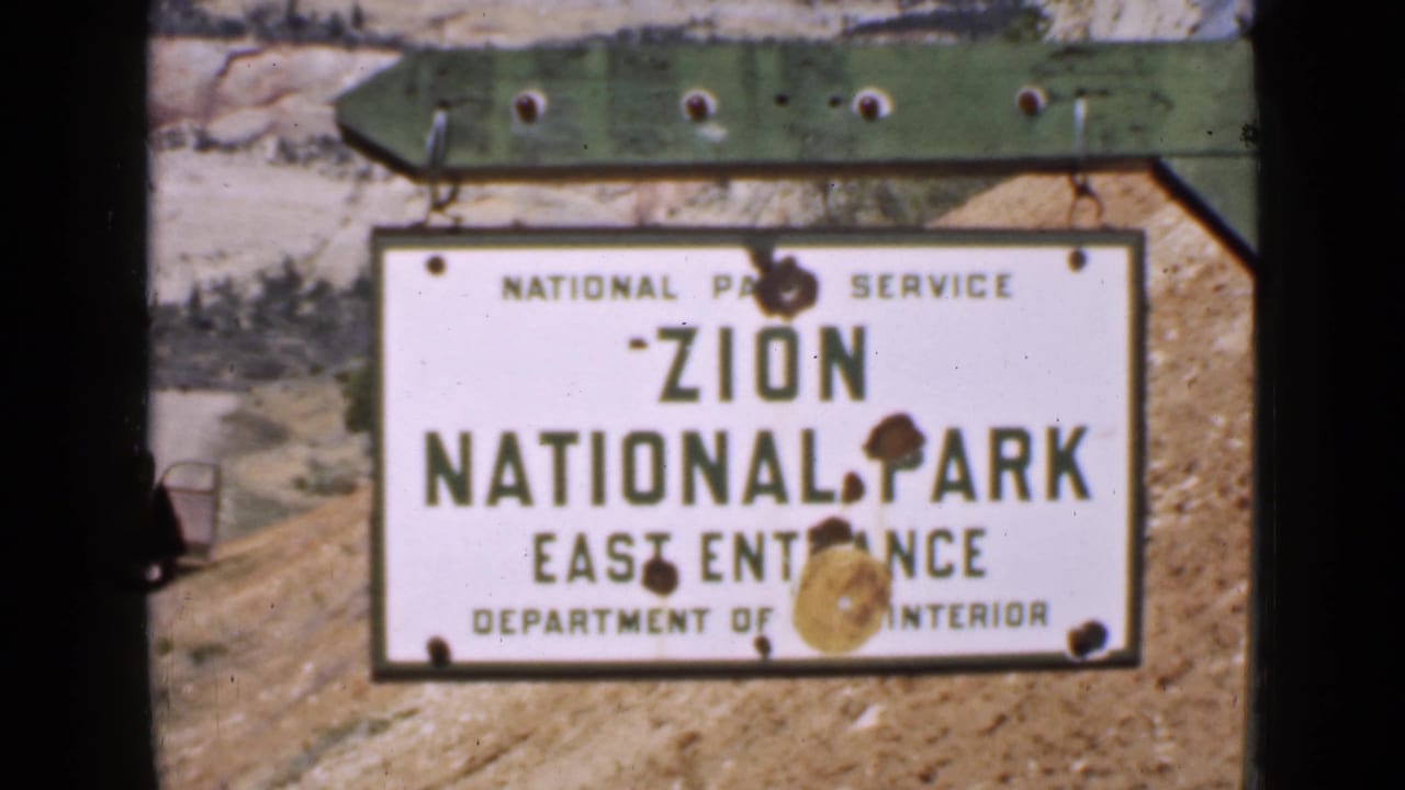 A close 1939 home-movie frame shows the Zion National Park East Entrance sign, with rust-like stains and dust marks across the image.