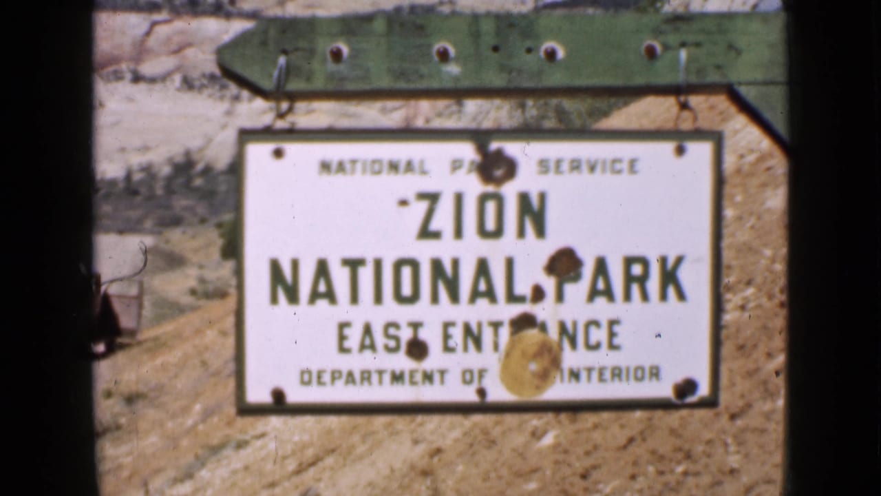 Final 8mm frame of the Zion National Park East Entrance sign in Utah, with a green support beam, dry roadside soil, and pale rock behind it.