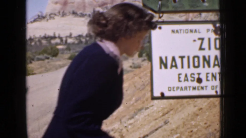 A woman in 1939 Utah moves past the camera with the Zion National Park East Entrance sign and desert hillside behind her.