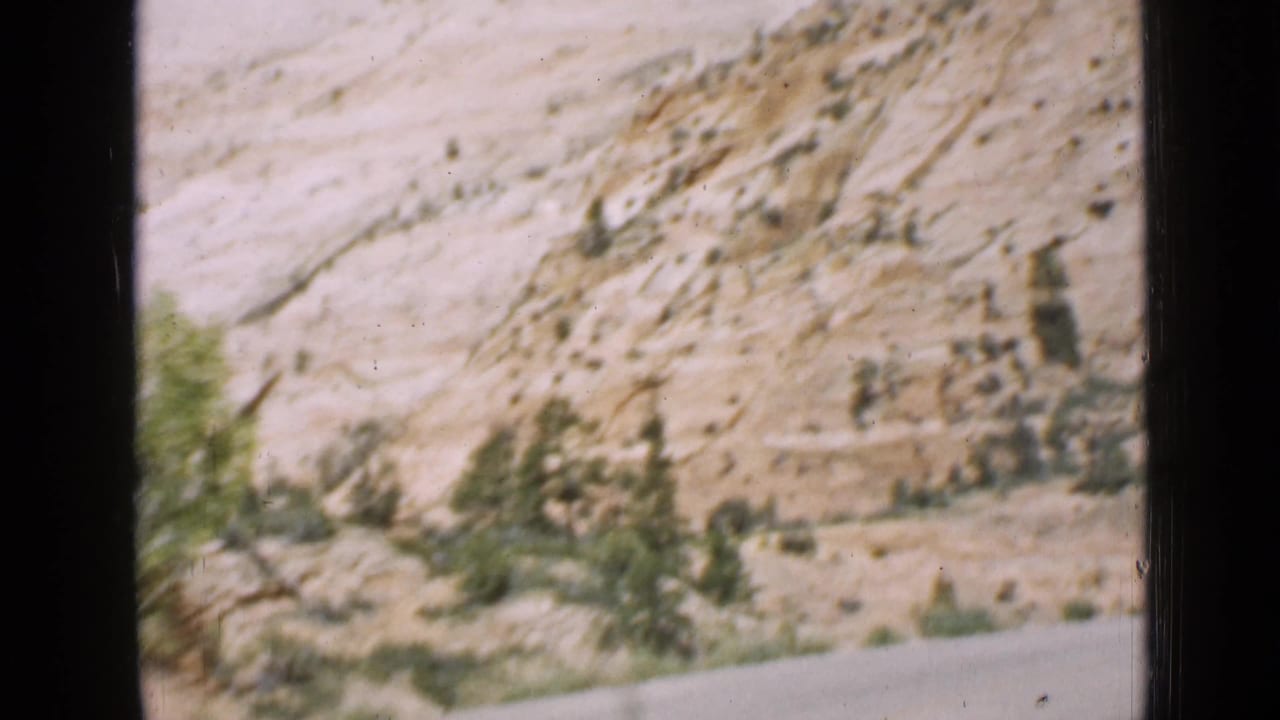 1939 8mm home-movie view from a moving car, showing a paved road curving past pale layered rock and sparse green shrubs in Utah.