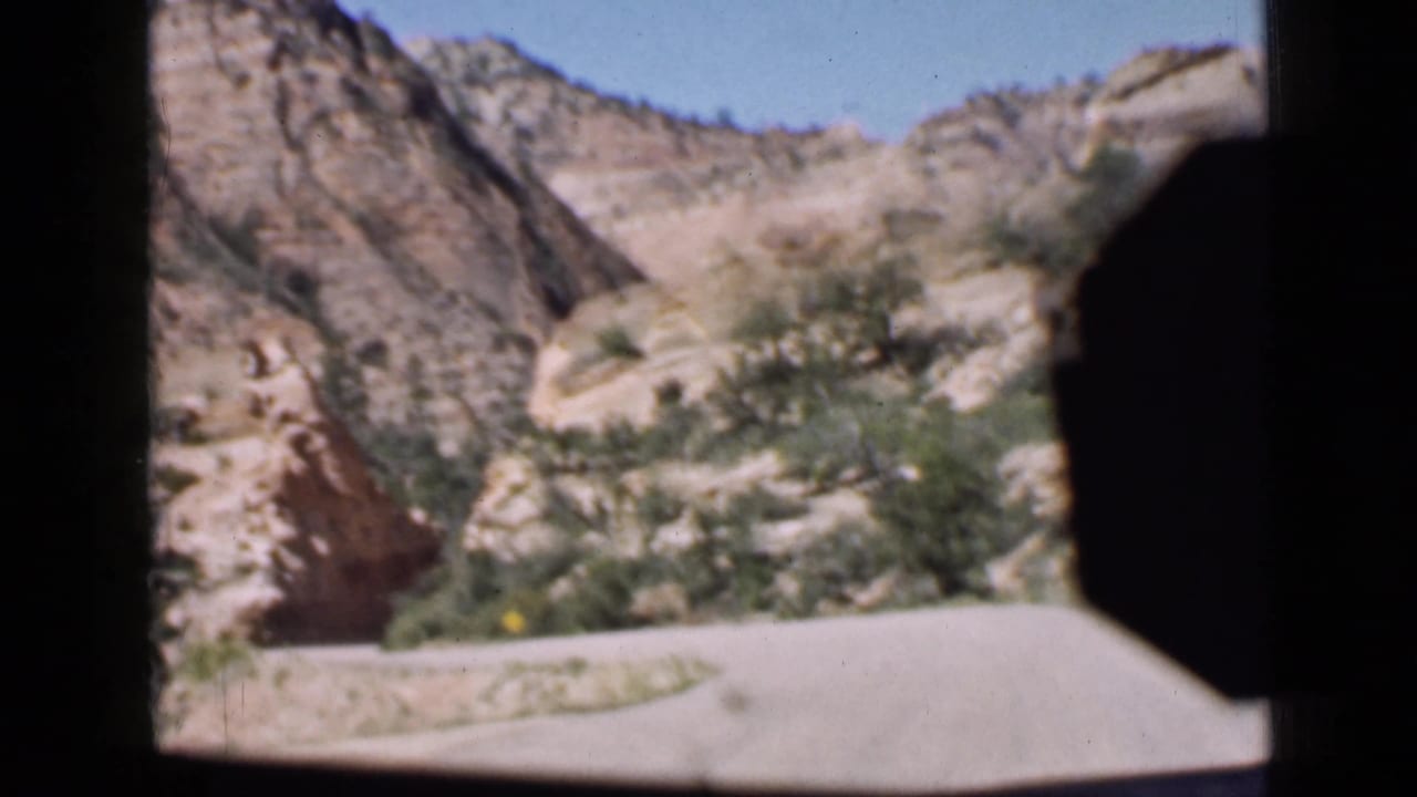 1939 Utah road view approaching a rocky canyon or tunnel area, with blue sky, roadside wall, shrubs, and a dark vehicle interior edge in frame.