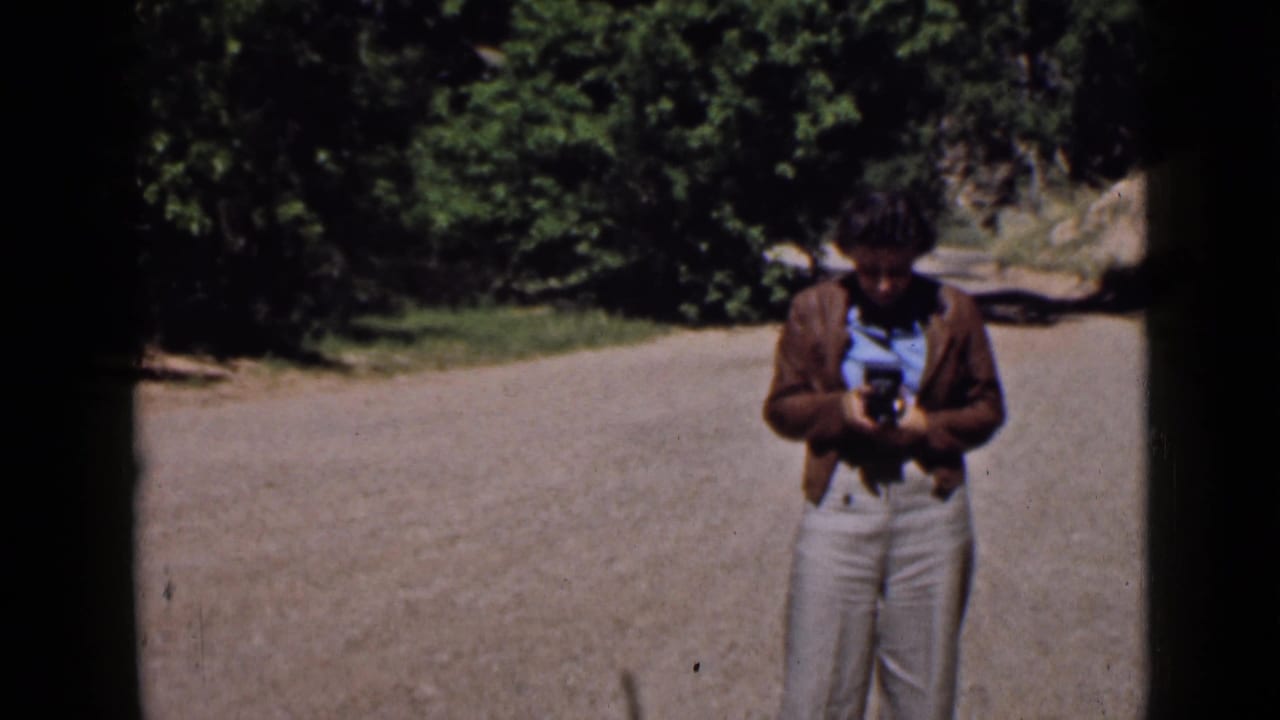 In 1939 Utah footage, an adult man stands on a dirt road adjusting a small camera, with trees and rocky ground behind him.