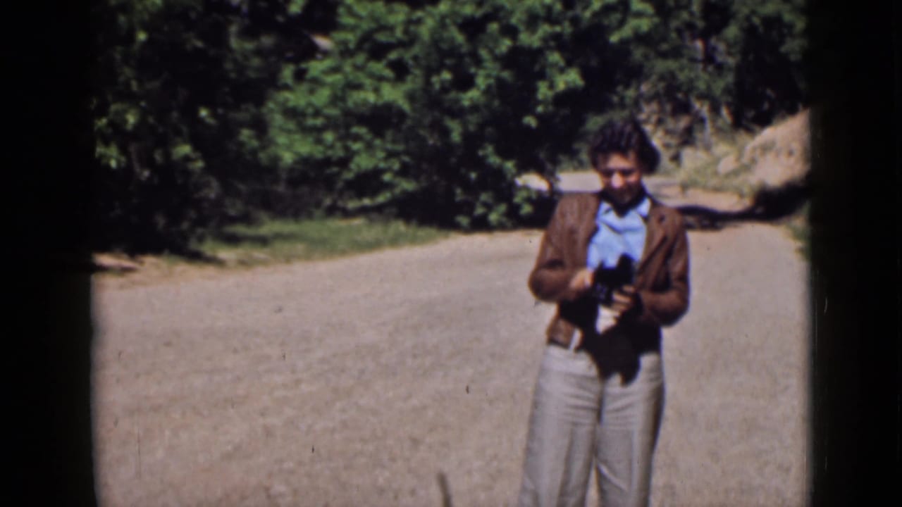 Adult man in a brown jacket stands on a Utah dirt road holding a camera, with roadside trees in the background.