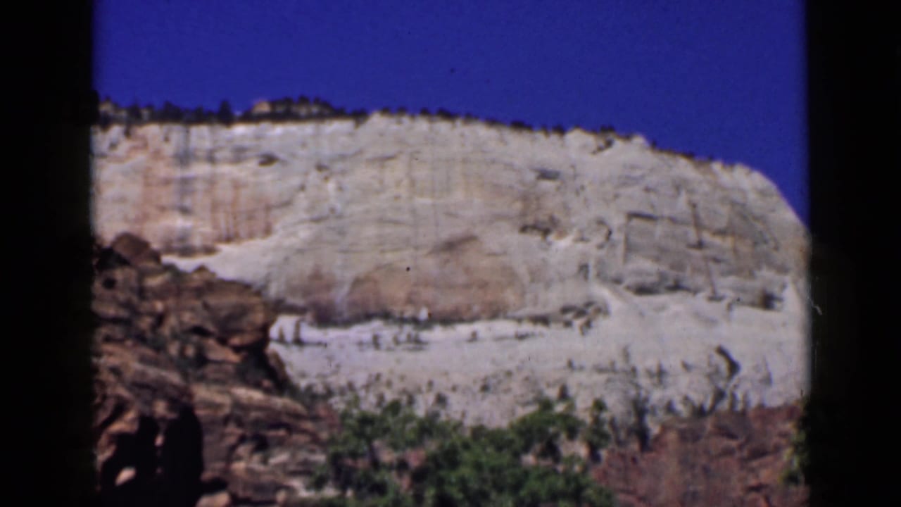 1939 Utah 8mm frame showing a pale sandstone-like cliff, blue sky, reddish foreground rock, and low green shrubs.