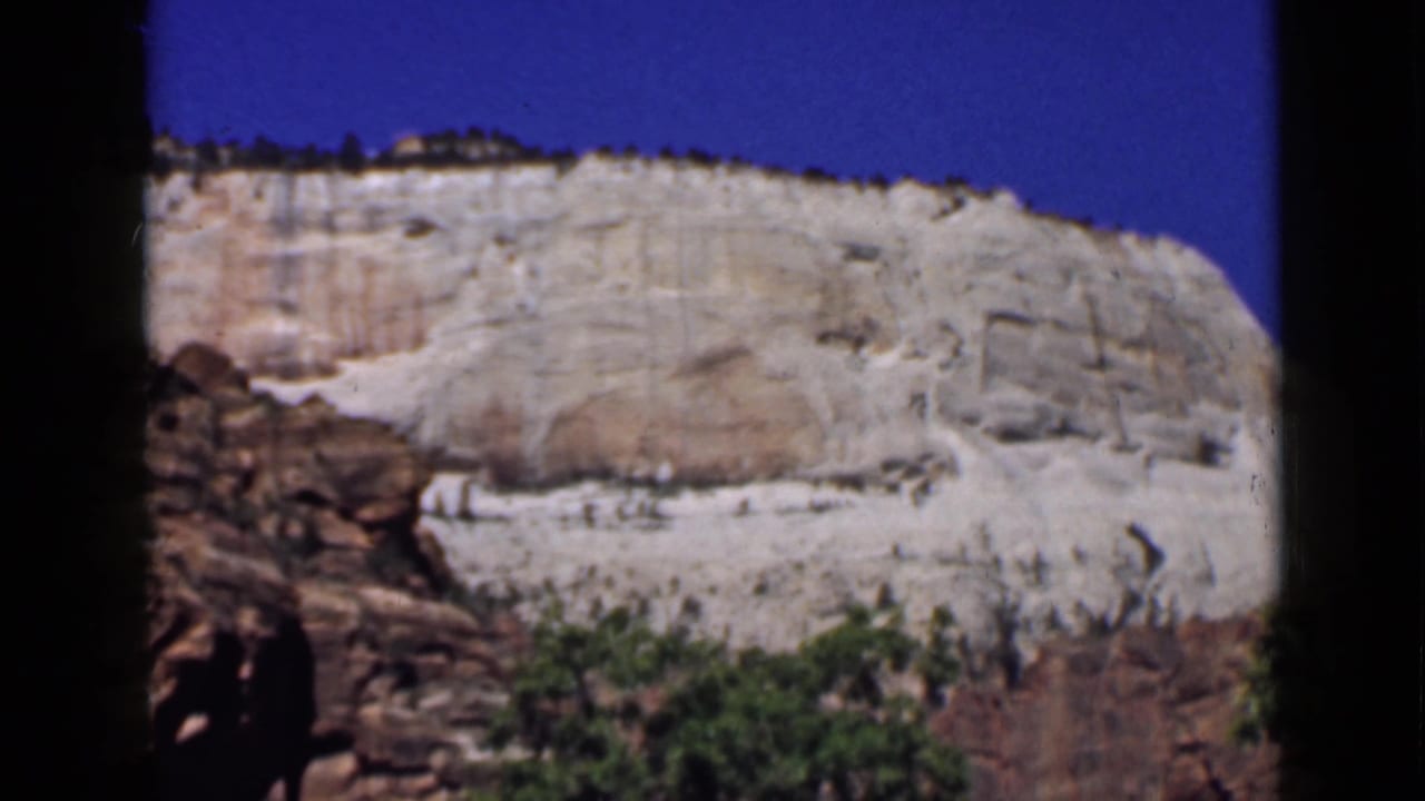 1939 Utah 8mm frame with a broad pale cliff face, clear blue sky, reddish rock at left, and green vegetation below.