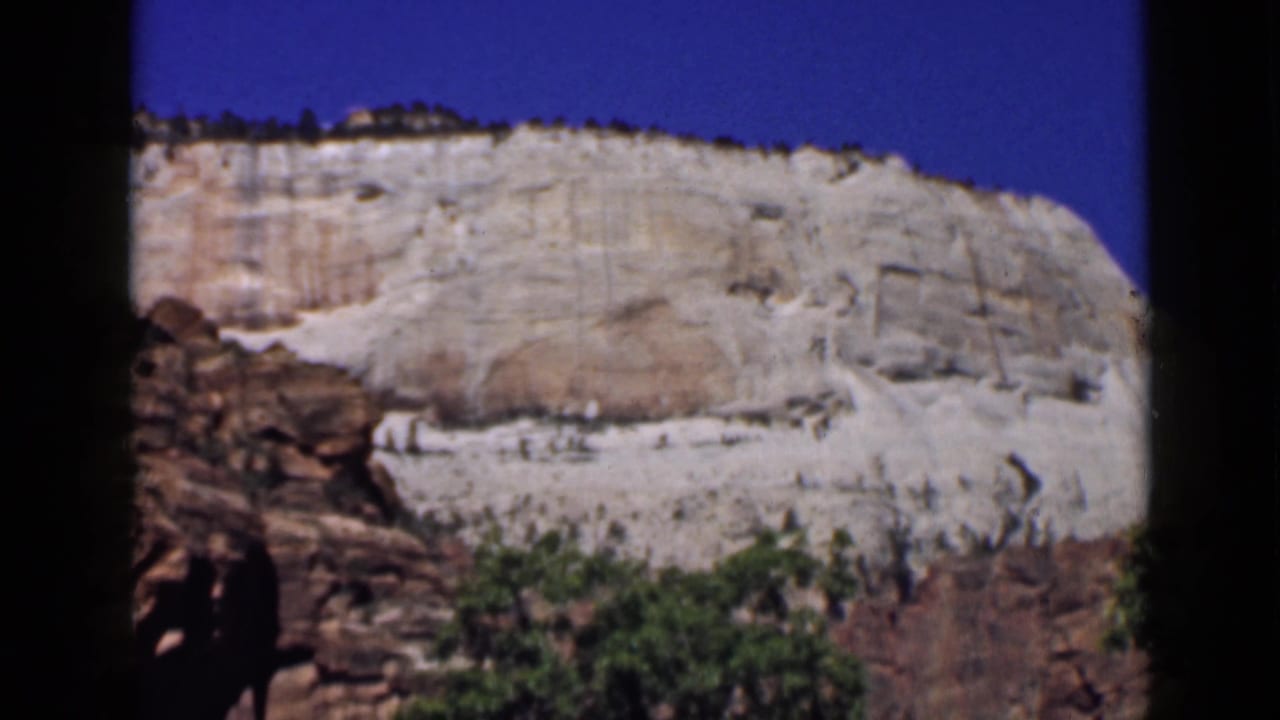 1939 Utah 8mm frame showing a sunlit pale rock formation above reddish rock and green shrubs under clear blue sky.