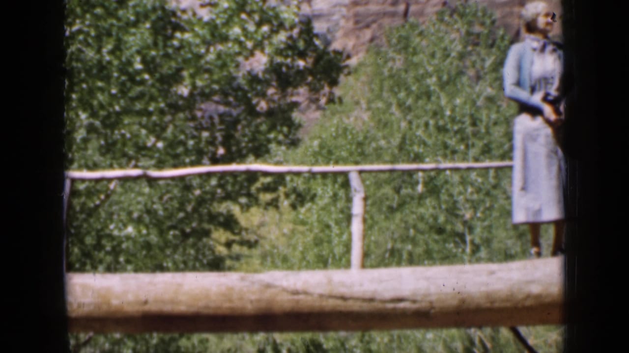 Utah 1939 frame showing a woman in a light skirt and cardigan on a rustic log bridge, with trees and a rocky ravine behind her.