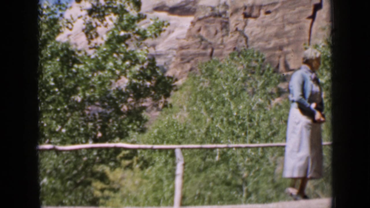 Utah 1939 home-movie frame with a woman near the end of a rustic log bridge, backed by green trees and a rocky ravine wall.