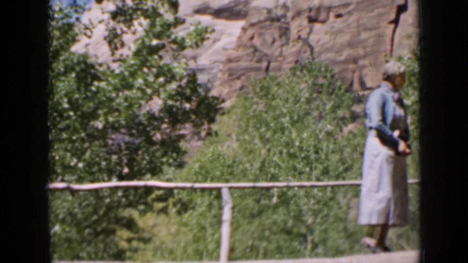Utah 1939 home-movie frame with a woman near the end of a rustic log bridge, backed by green trees and a rocky ravine wall.