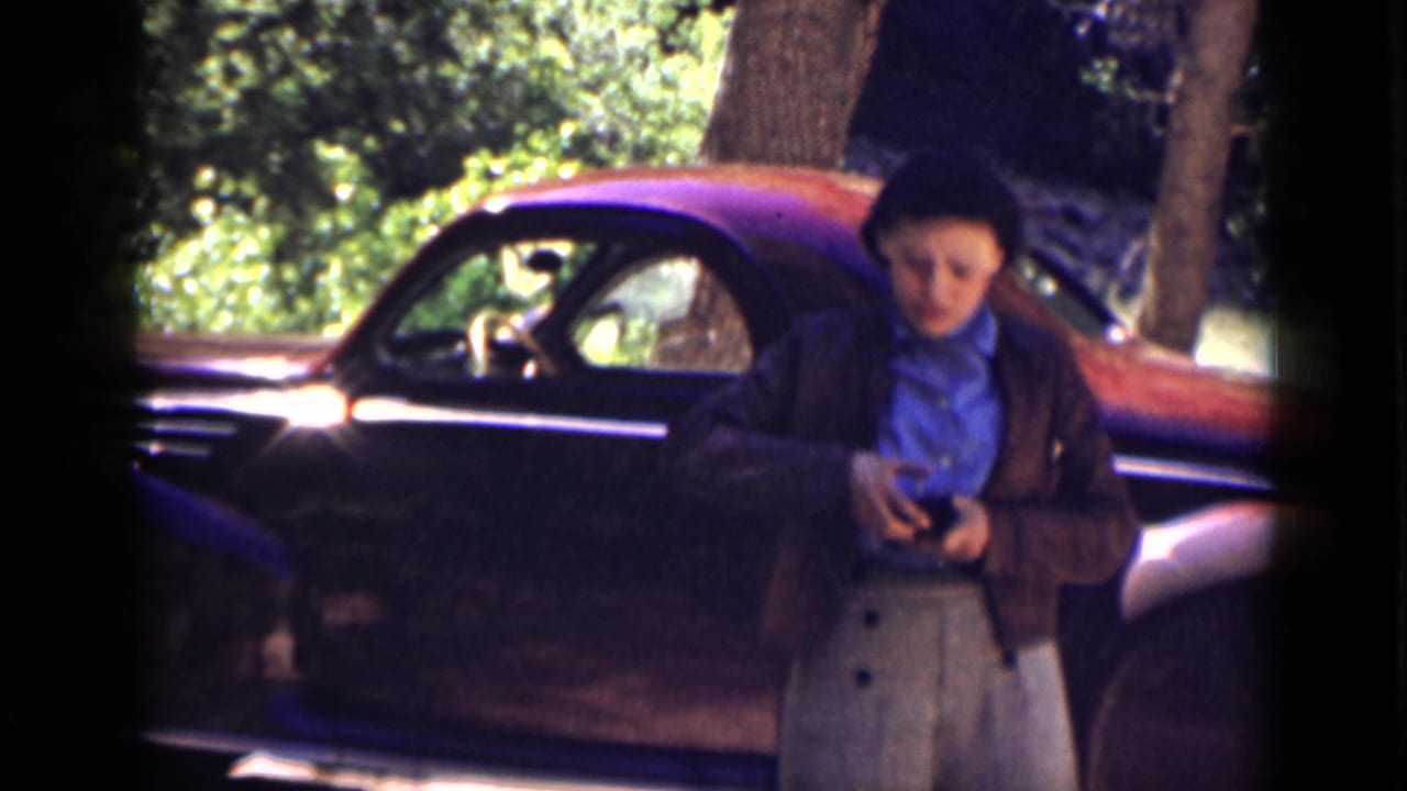 In Utah in 1939, a woman in a blue shirt and dark jacket stands beside a classic car, holding a small object.