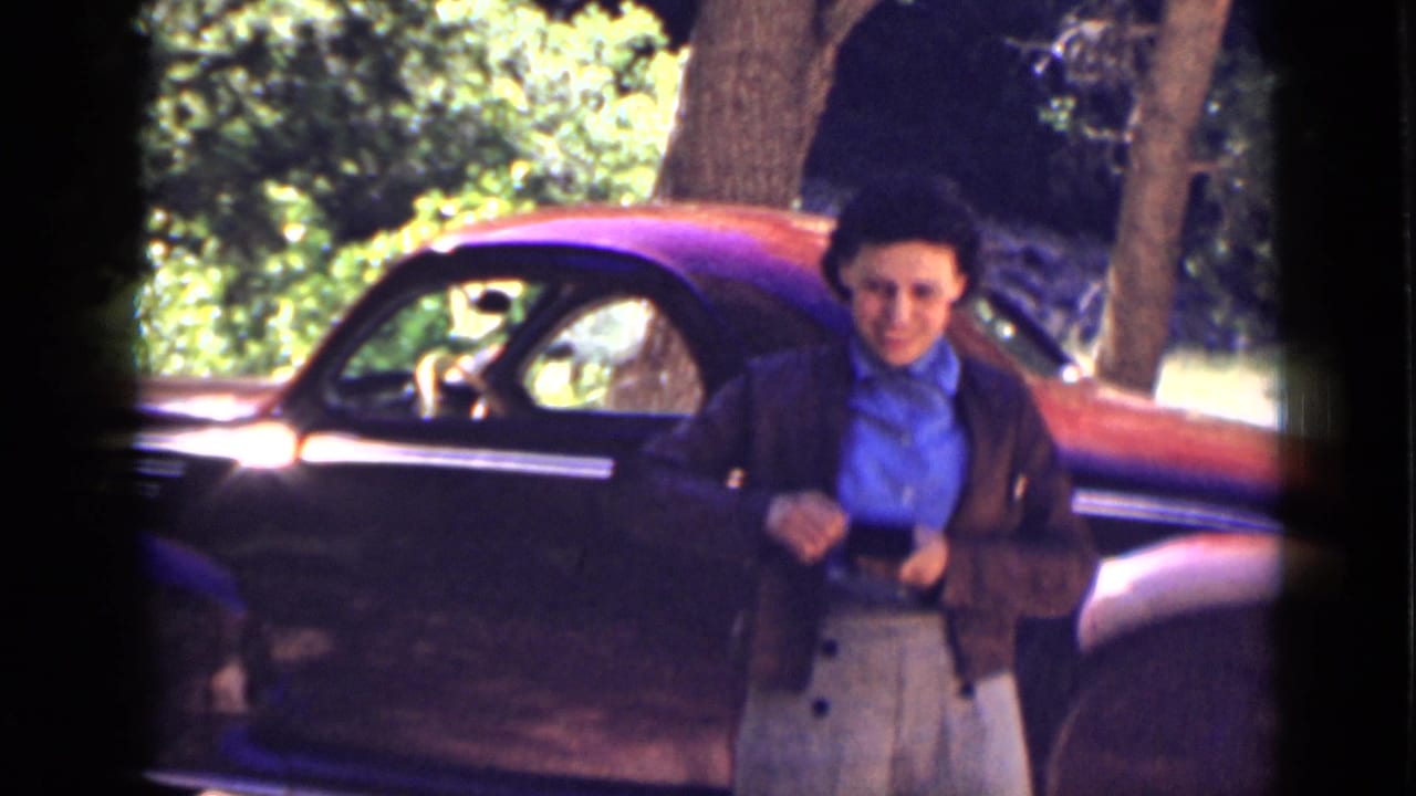 In 1939 Utah home-movie footage, an elderly woman stands before a red car beside trees under harsh sun and lens flare.
