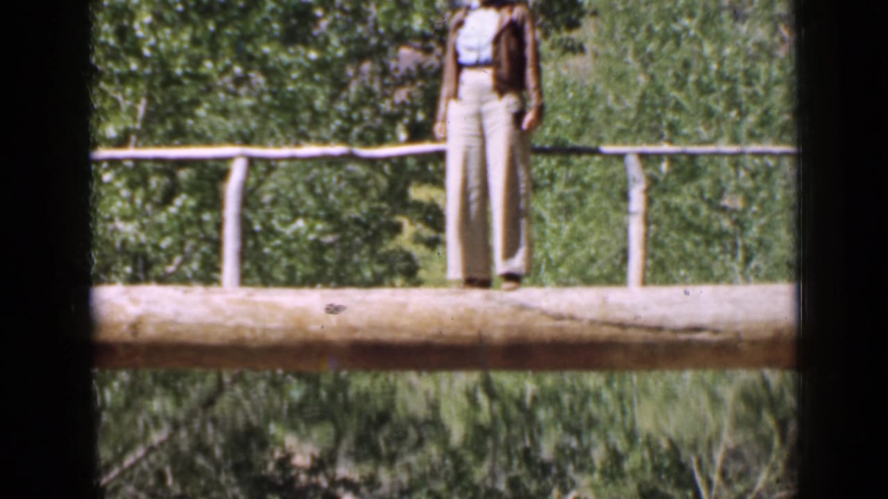 1939 Utah home-movie frame of a woman in light clothing standing on a rustic wooden bridge above dense foliage.