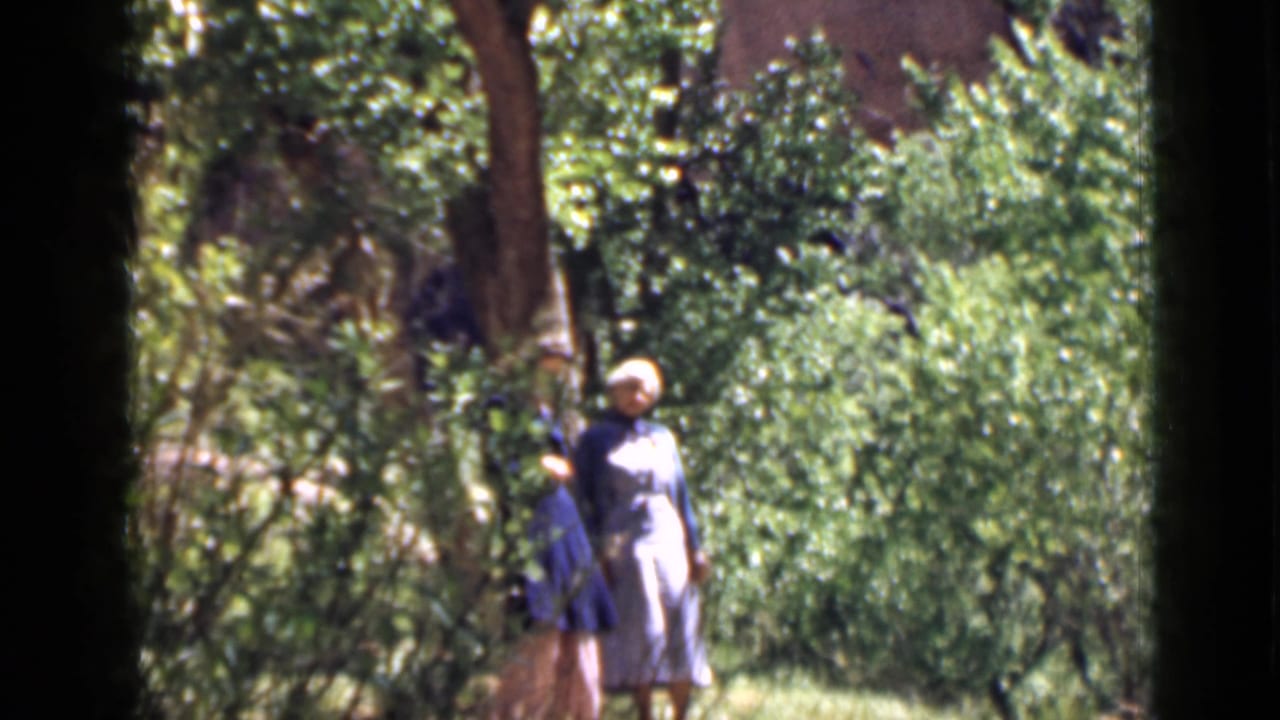 Two women appear near dense green foliage in 1939 Utah home-movie footage, with trees and rock walls around the scene.
