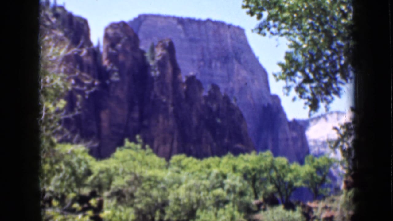 1939 Utah 8mm frame showing a broad rock face, darker vertical spires, green trees, and a branch-framed scenic view.