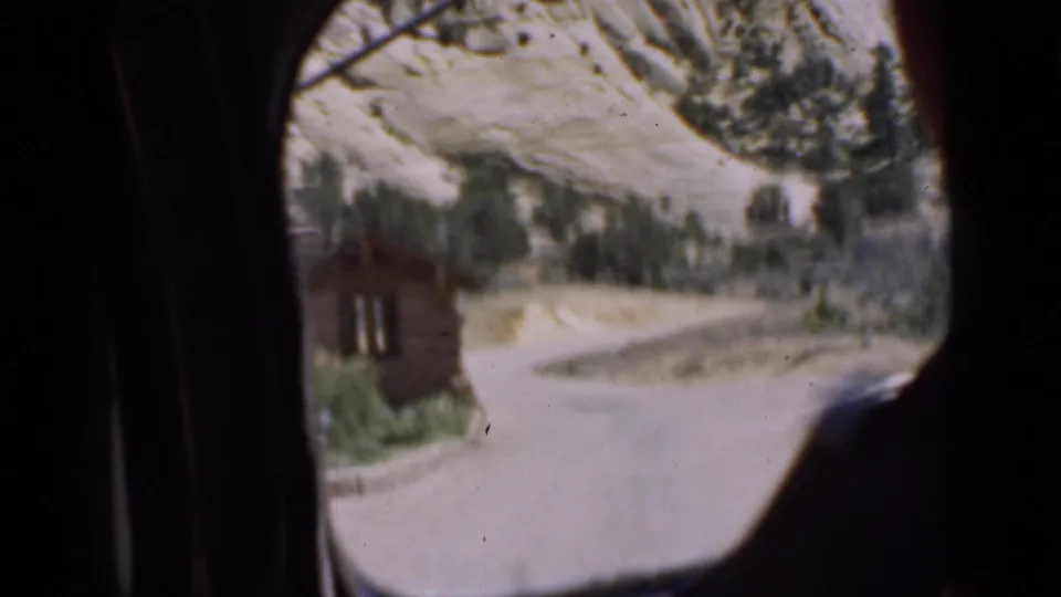 1939 Utah travel frame seen through a car window, with a small wooden cabin near a winding road and rocky hillside beyond.