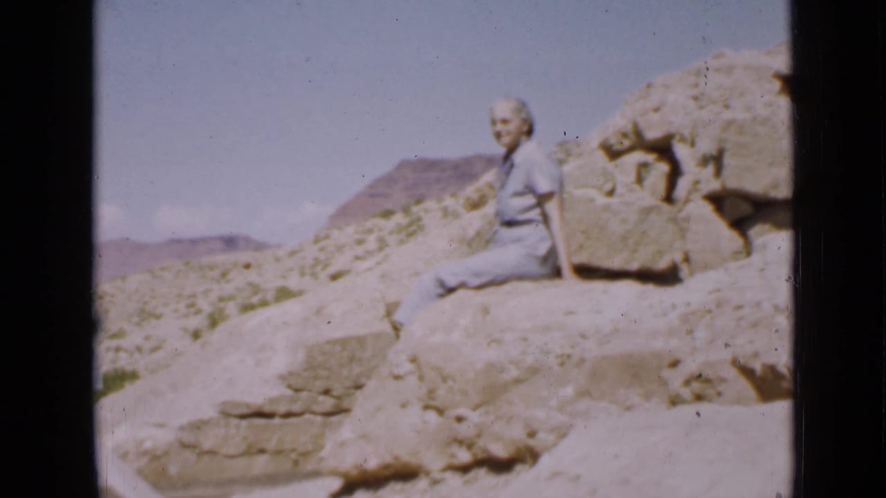 In 1939 Utah home-movie footage, a woman sits on pale desert rocks with arid hills and sparse vegetation behind her.