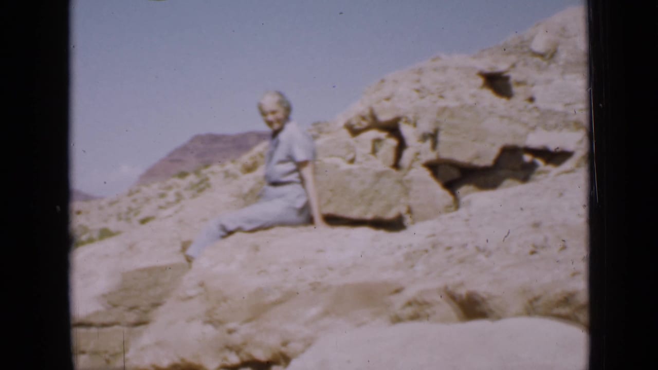 A softly focused 1939 Utah frame shows the woman sitting beside large boulders with sunlit desert ground and hills behind her.
