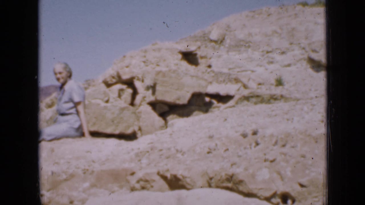 1939 Utah home-movie frame of a woman in a blue dress seated on rocky desert outcrop beneath a clear sky.