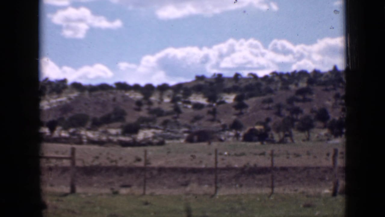 1939 Utah home-movie frame showing a wire fence, open ground, shrub-covered hills, and bright clouds, framed by dark film edges.