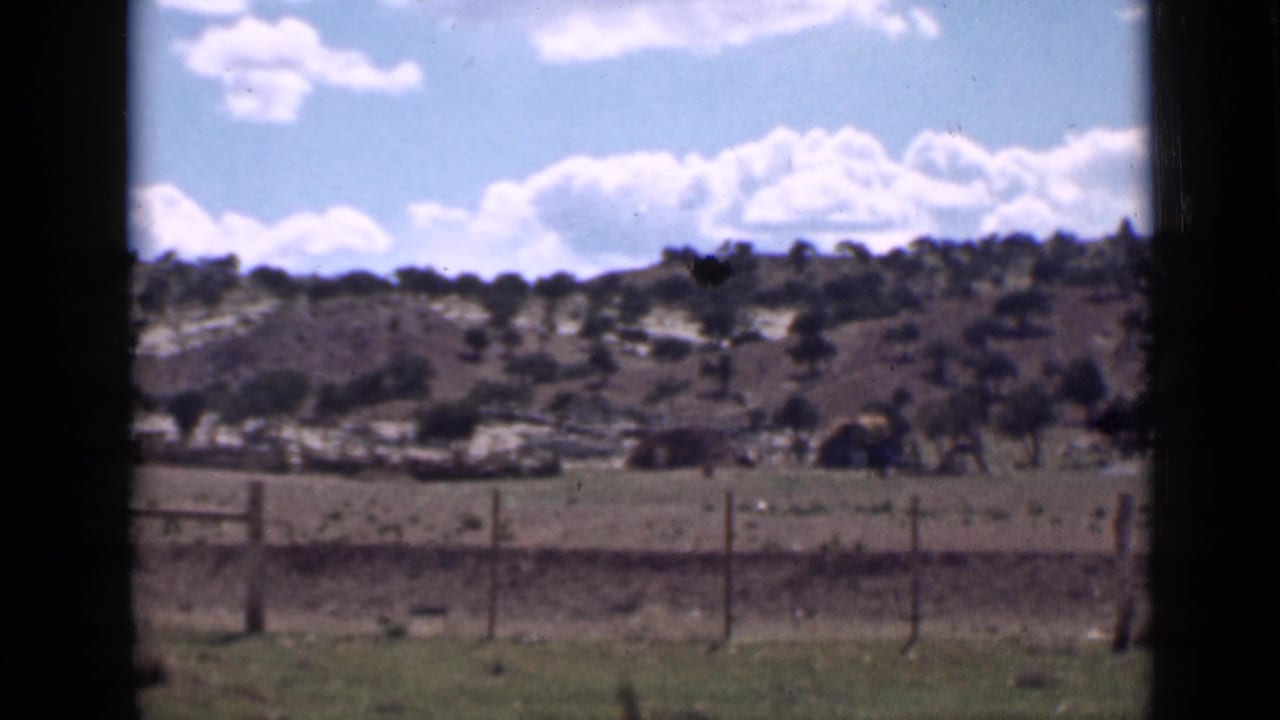 1939 Utah home-movie frame showing dry prairie, a wooden fence, scattered trees, low hills, and a bright clouded sky.