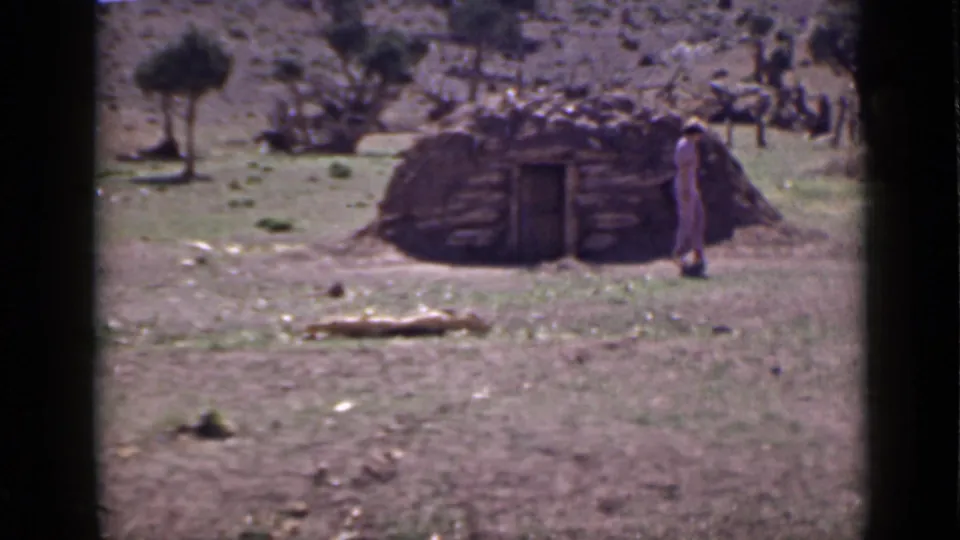 1939 Utah home-movie frame with a rustic hut, open doorway, lone person, dry foreground, and sparse trees on the hillside.