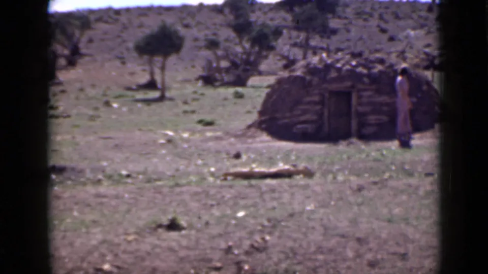 1939 Utah frame showing a lone figure beside a small hut, with dry ground, scattered brush, and rocky hills in the background.